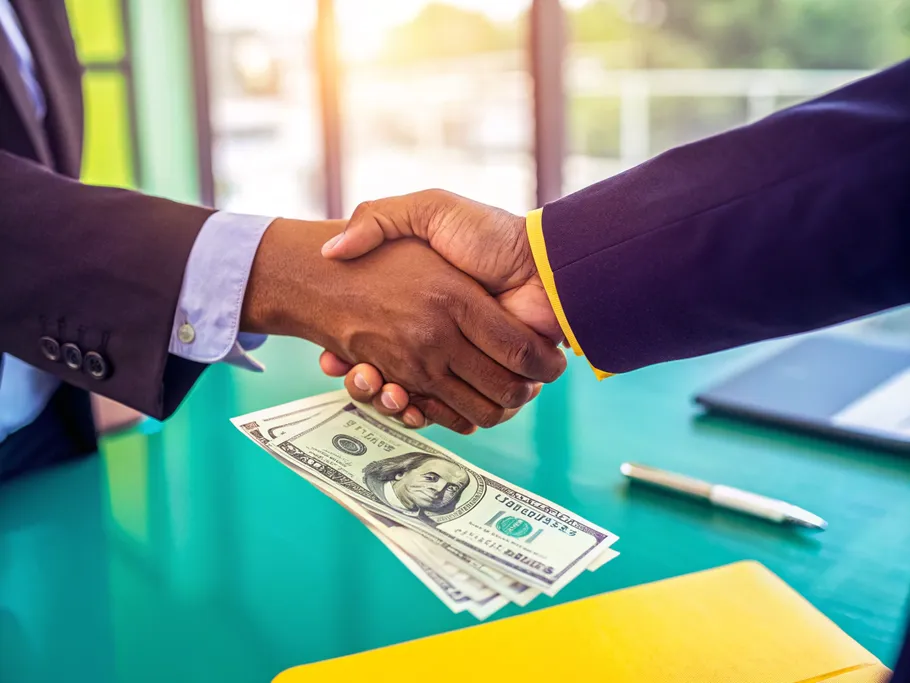 Two professionals shaking hands over a table, one handing over cash to the other in a bright office setting